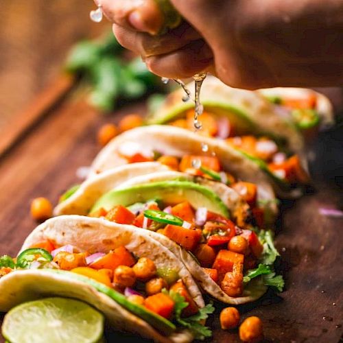 A hand squeezes lime over colorful tacos filled with chickpeas, vegetables, and fresh herbs, resting on a wooden board.