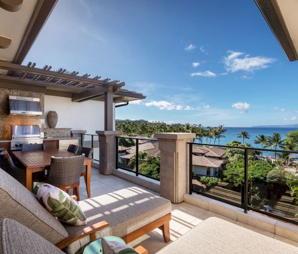The image features a stylish balcony with a dining area, overlooking a scenic beach and lush greenery under a blue sky.