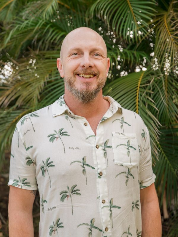 A person wearing a palm tree-patterned shirt stands in front of lush green foliage, smiling at the camera.