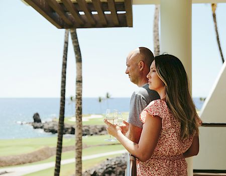A couple holding wine glasses enjoys a scenic ocean view from a balcony, surrounded by palm trees and clear skies.