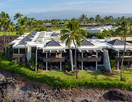 Luxurious beachfront villa with white modern architecture, palm trees, and lava rock coastline under a sunny blue sky.
