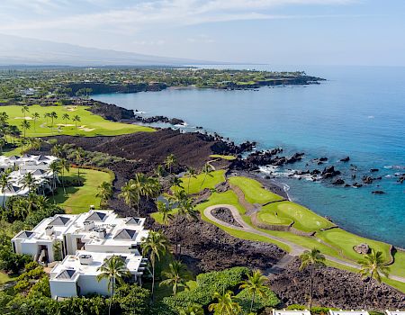 A tropical coastal resort with white buildings, lush green lawns, palm trees, rocky shoreline, and turquoise ocean under a blue sky, coastal golf course scenery.