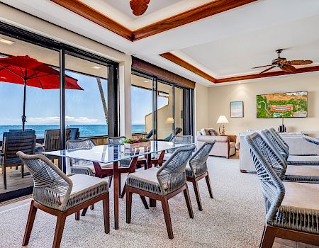 A bright coastal living space with a glass wall to a patio, wicker chairs around a glass table, red umbrella, and ocean view.