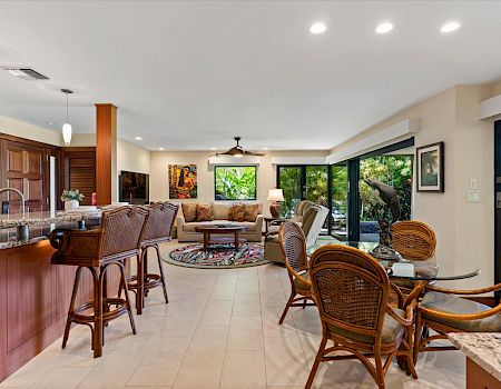 A bright, open-concept living area with a kitchen island and bar stools, a cozy seating lounge, and large windows/doors bringing in greenery.