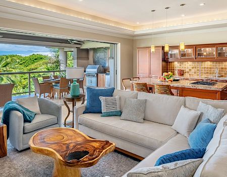 A bright, open living room with a curved white sofa, blue cushions, a wood coffee table, and an adjacent airy kitchen and outdoor dining area with greenery.