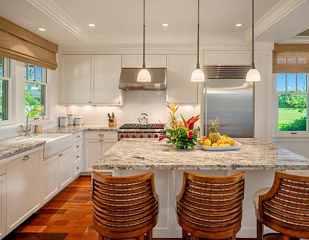 A bright, spacious kitchen with white cabinets, marble island, pendant lights, stainless steel appliances, and wooden bar stools by the island.