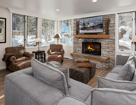 Cozy living room with a stone fireplace, gray couches, wooden coffee table, and large windows showing snowy outdoors, warm lighting, and artwork on the wall.