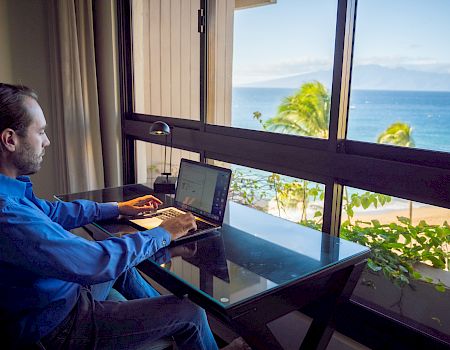 A man with a beard works on a laptop at a glass balcony desk, overlooking a sunny ocean view with palm trees.