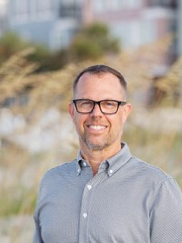 A smiling man with glasses in a light blue button-up shirt stands outdoors with a blurred grassy and urban background.