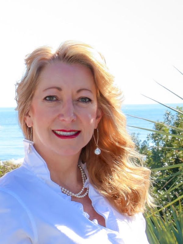 A smiling woman with blonde hair, wearing a white blouse and pearl necklace, outdoors by the sea with blue sky and greenery behind her.