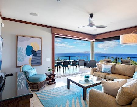 A bright, modern living area with a beige sectional, blue accents, a coffee table, and a view of the ocean through large windows and a balcony.
